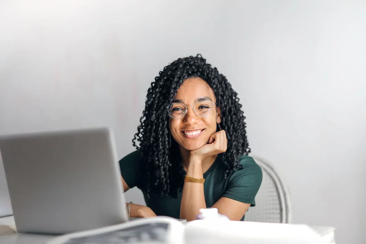 A woman sitting at a table with an open laptop, smiling, a notebook and papers in front of her