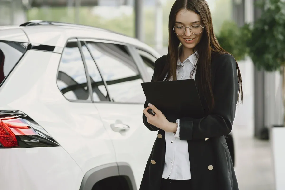 A woman standing beside a white SUV outdoors holding a closed document folder, ready for a dealership appointment