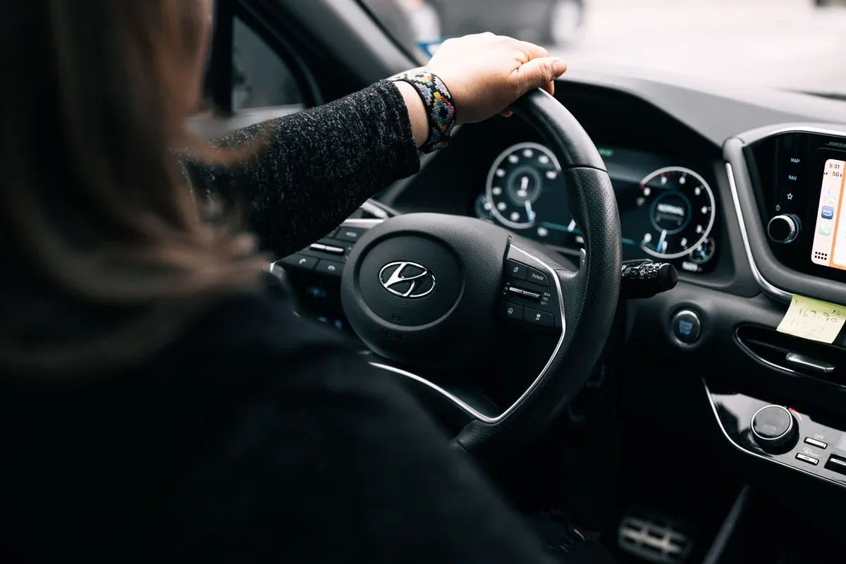 View over the shoulder of a woman driving, one hand on the steering wheel, dashboard and controls lit ahead of her