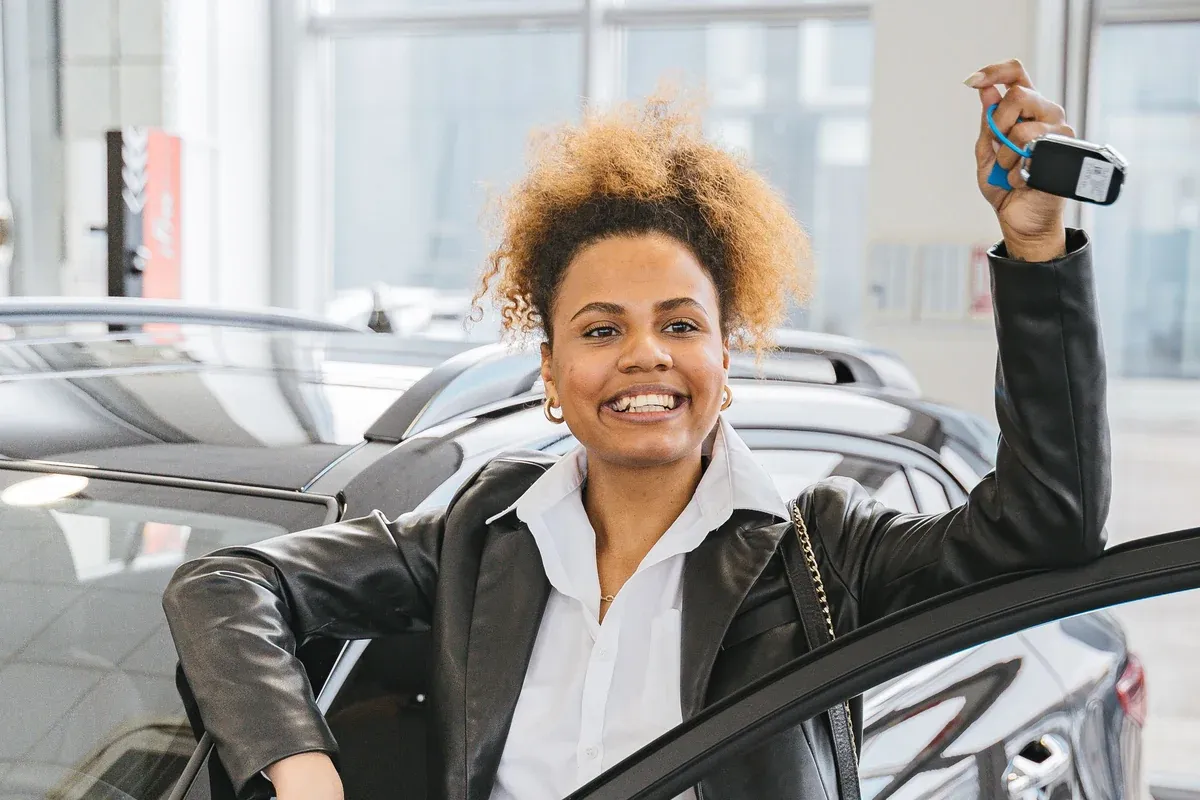 A woman standing at the open door of the car she has just bought, holding up the key in a dealership showroom