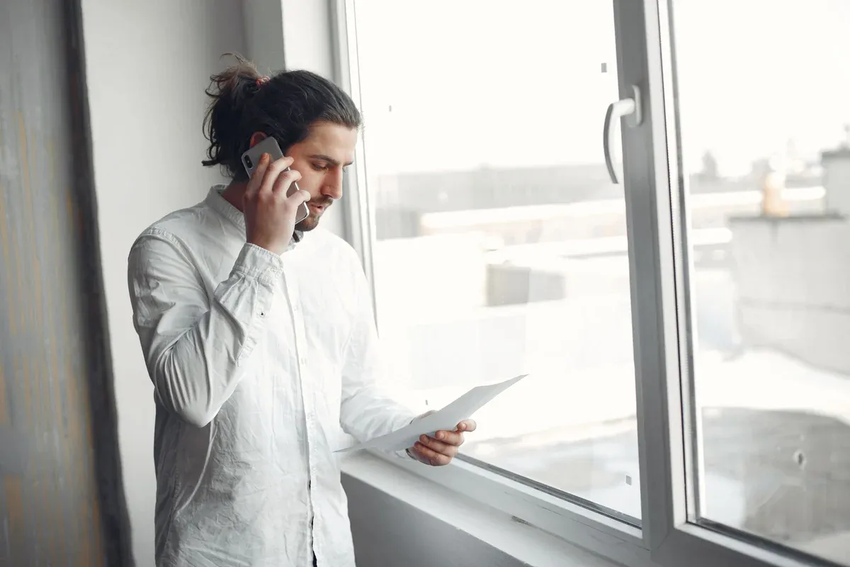 A man standing by a window holding a printed statement in one hand while talking on his mobile phone
