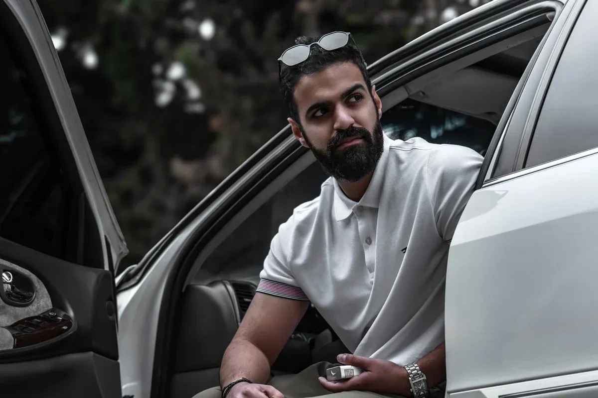 A young man sitting in the open driver's door of a used sedan outdoors, holding the key after buying the car himself