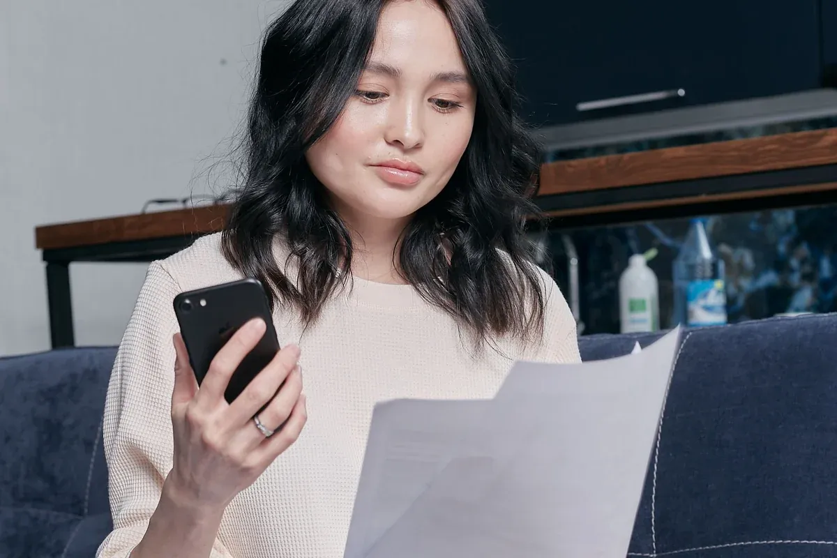 A woman sitting at home reading a document, with a phone in one hand and envelopes, paperwork and a laptop spread out beside her