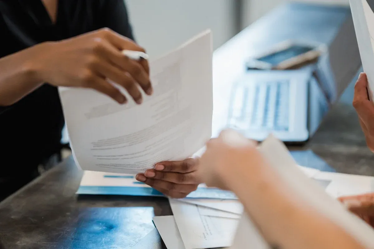 Close-up of two people at a desk passing a printed document between them, with envelopes, a calculator and paperwork on the table