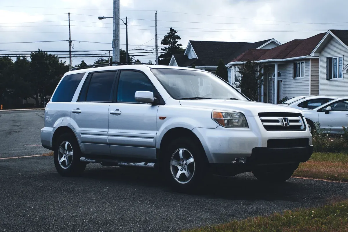 A white used Honda Pilot SUV parked on a residential street outside a suburban house on an overcast day