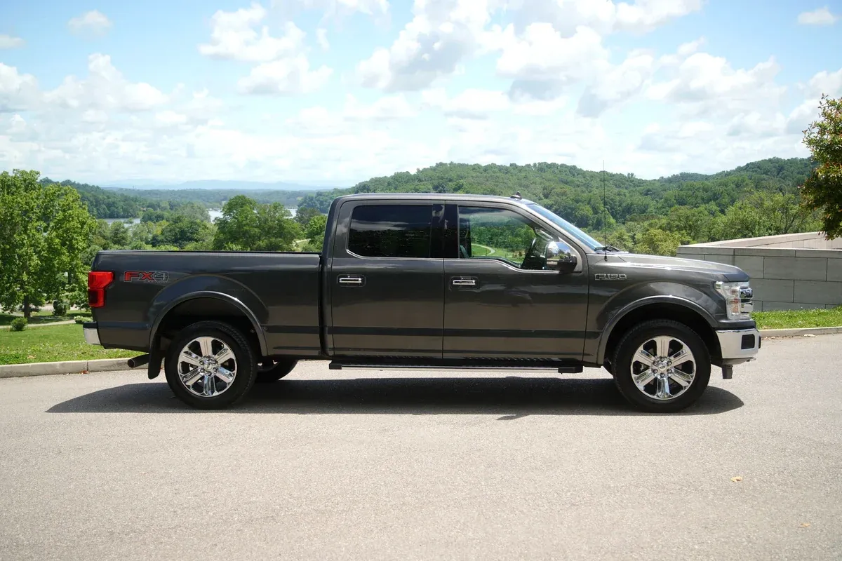 A grey used Ford F-150 crew cab pickup truck photographed in profile on a paved lot with green hills behind it